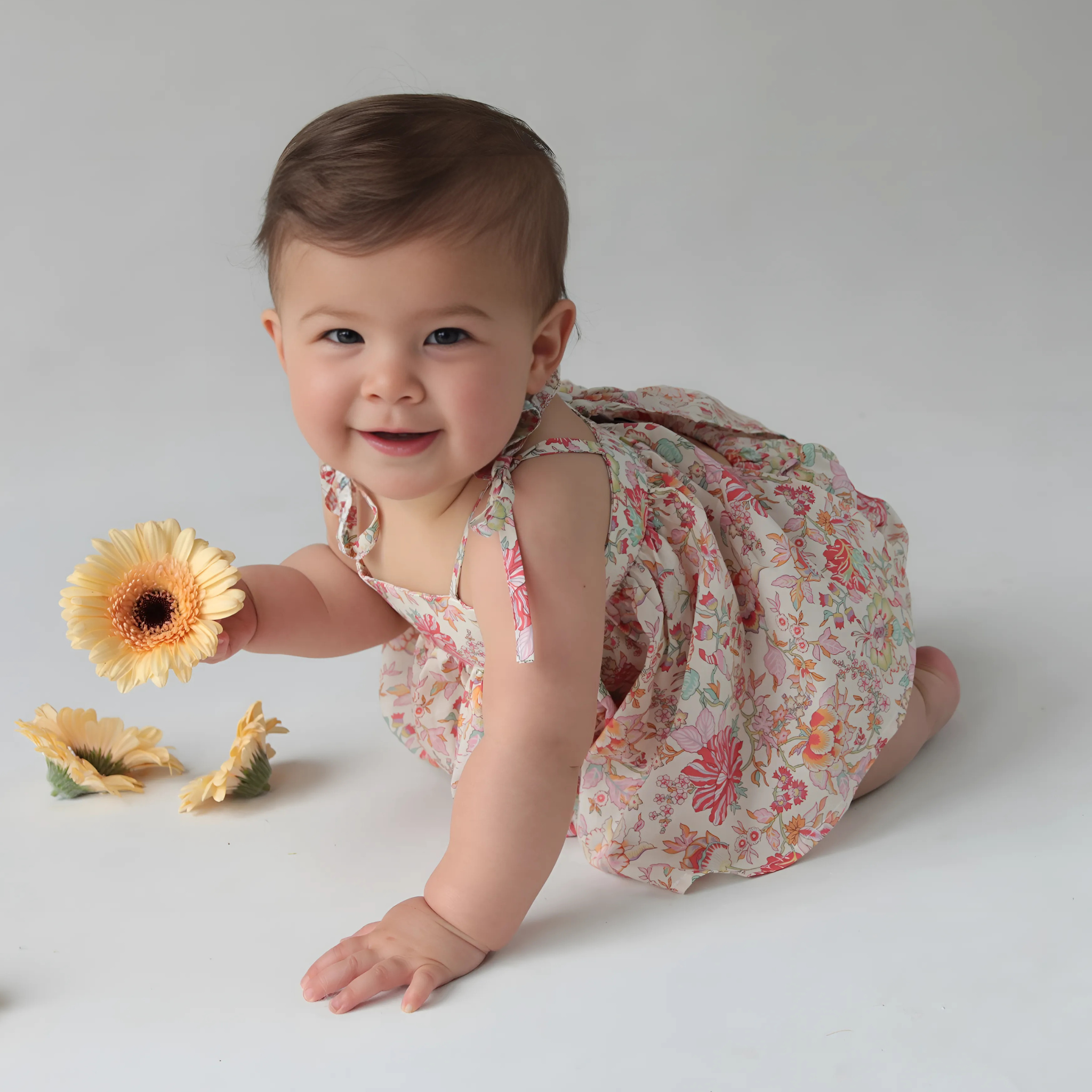 Baby in a floral dress holding a flower on a plain background