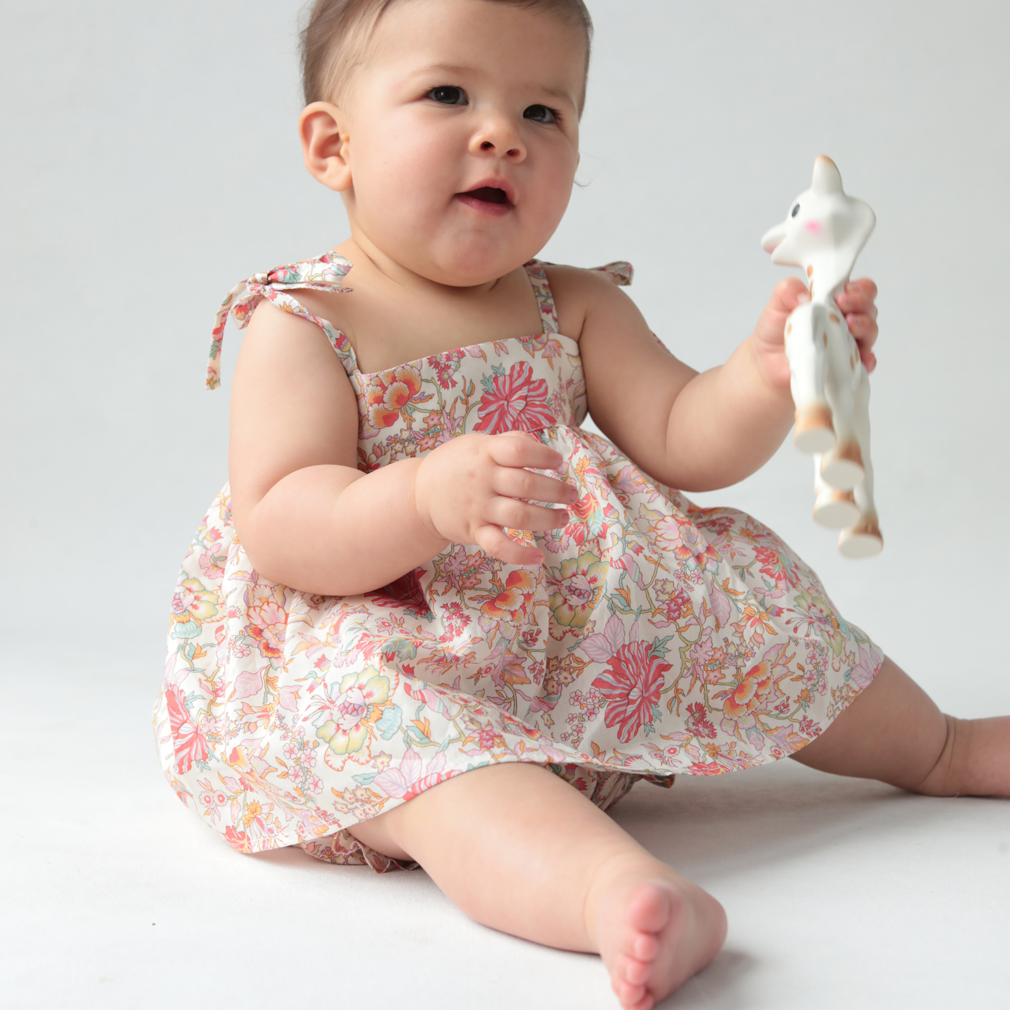 Baby in a floral blouse holding a toy against a plain background