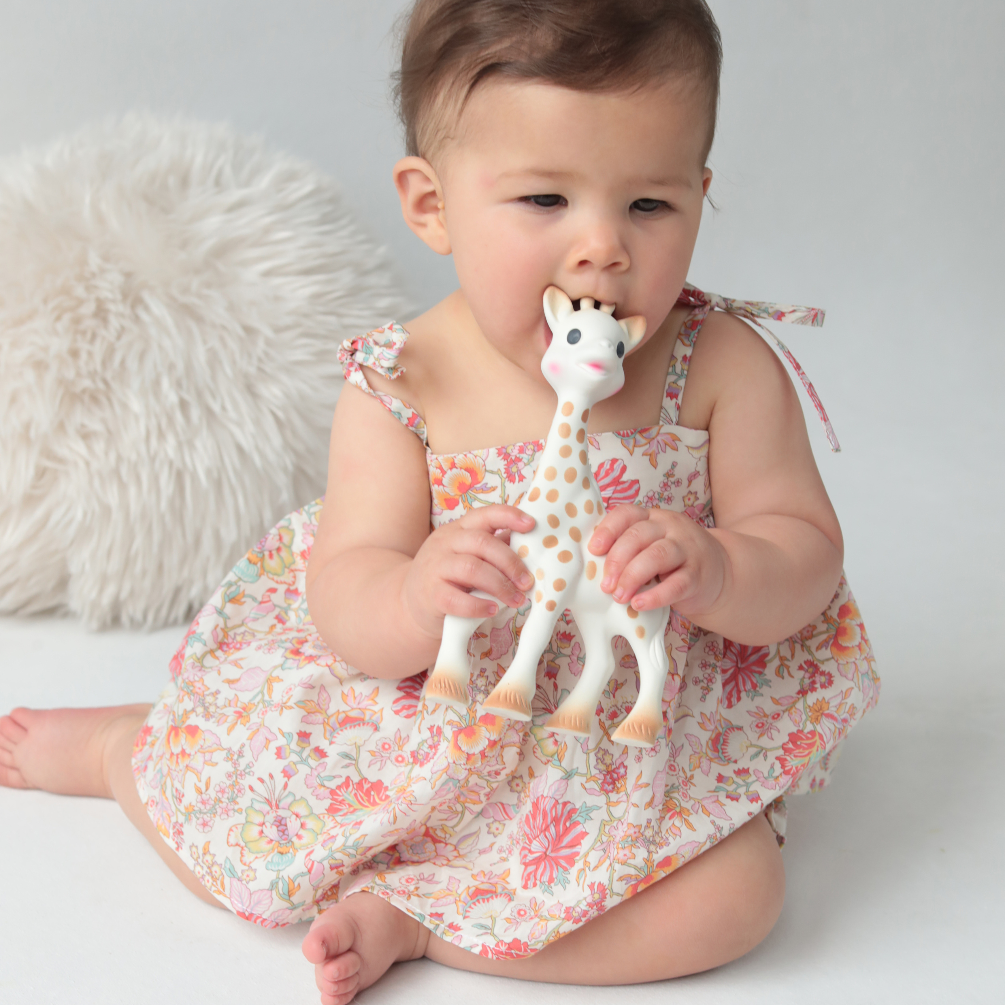Baby in a floral blouse holding a giraffe-shaped toy against a neutral background