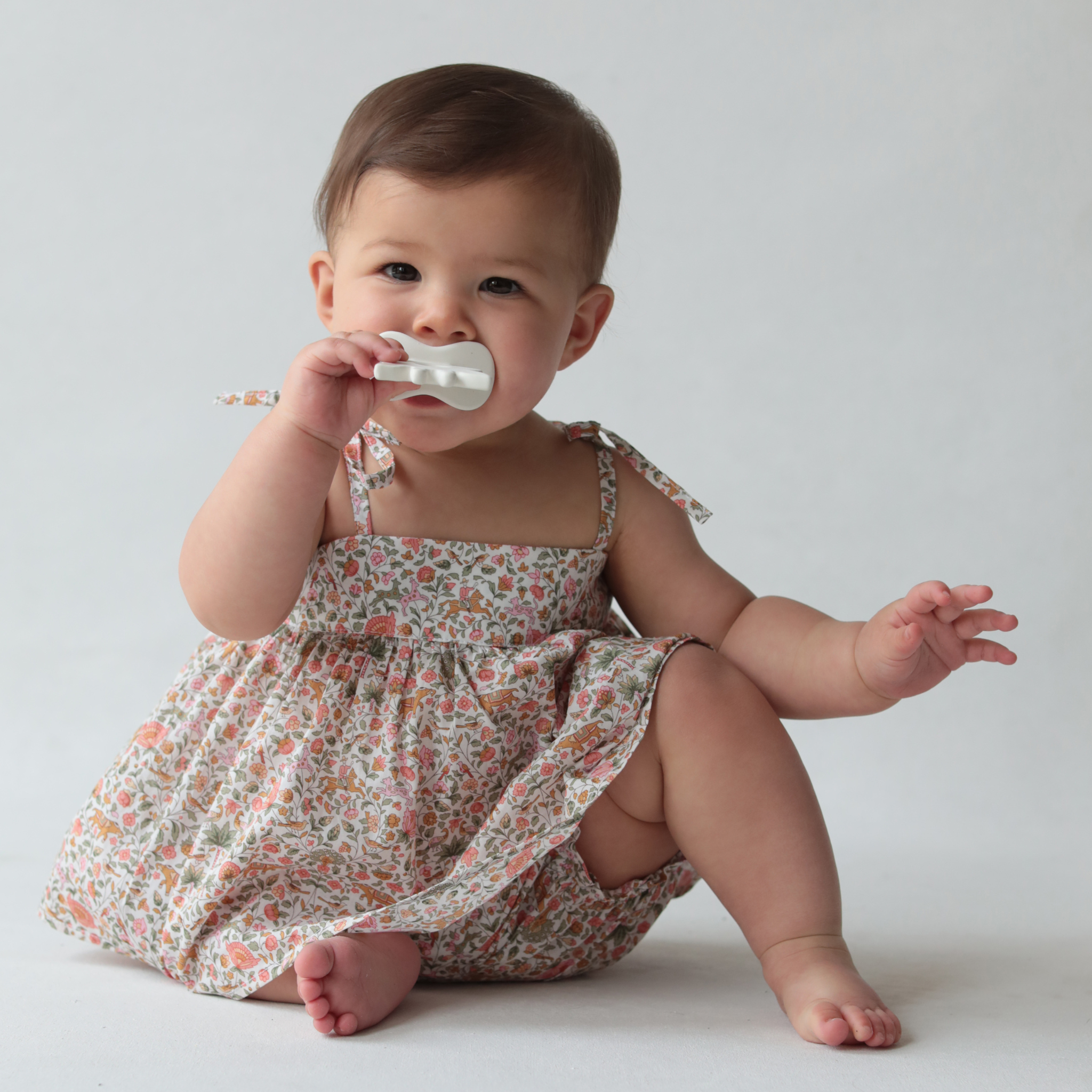 Baby in a floral dress sitting on a white surface with a plain background