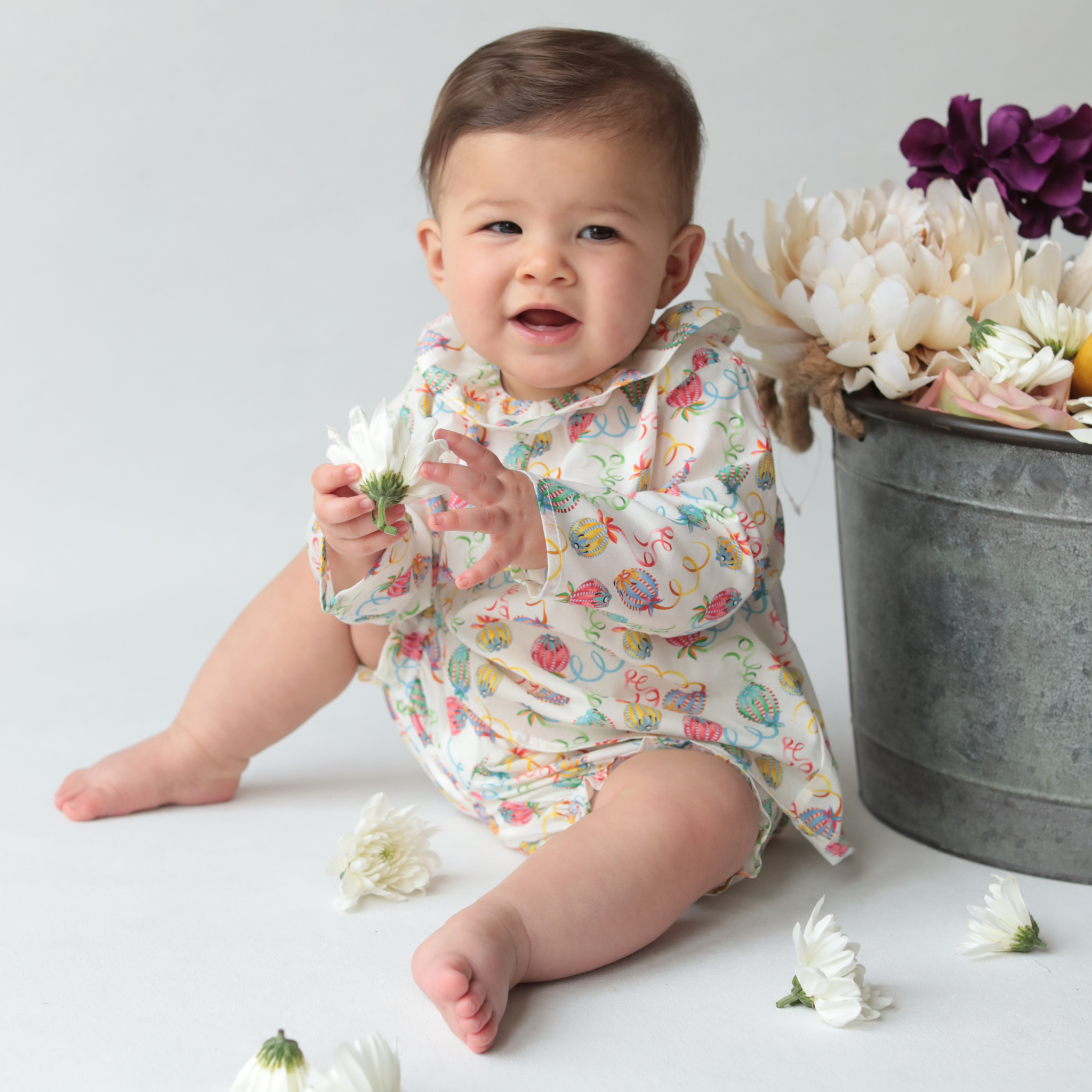 Baby in a floral blouse and bloomer sitting on a white surface with flowers around