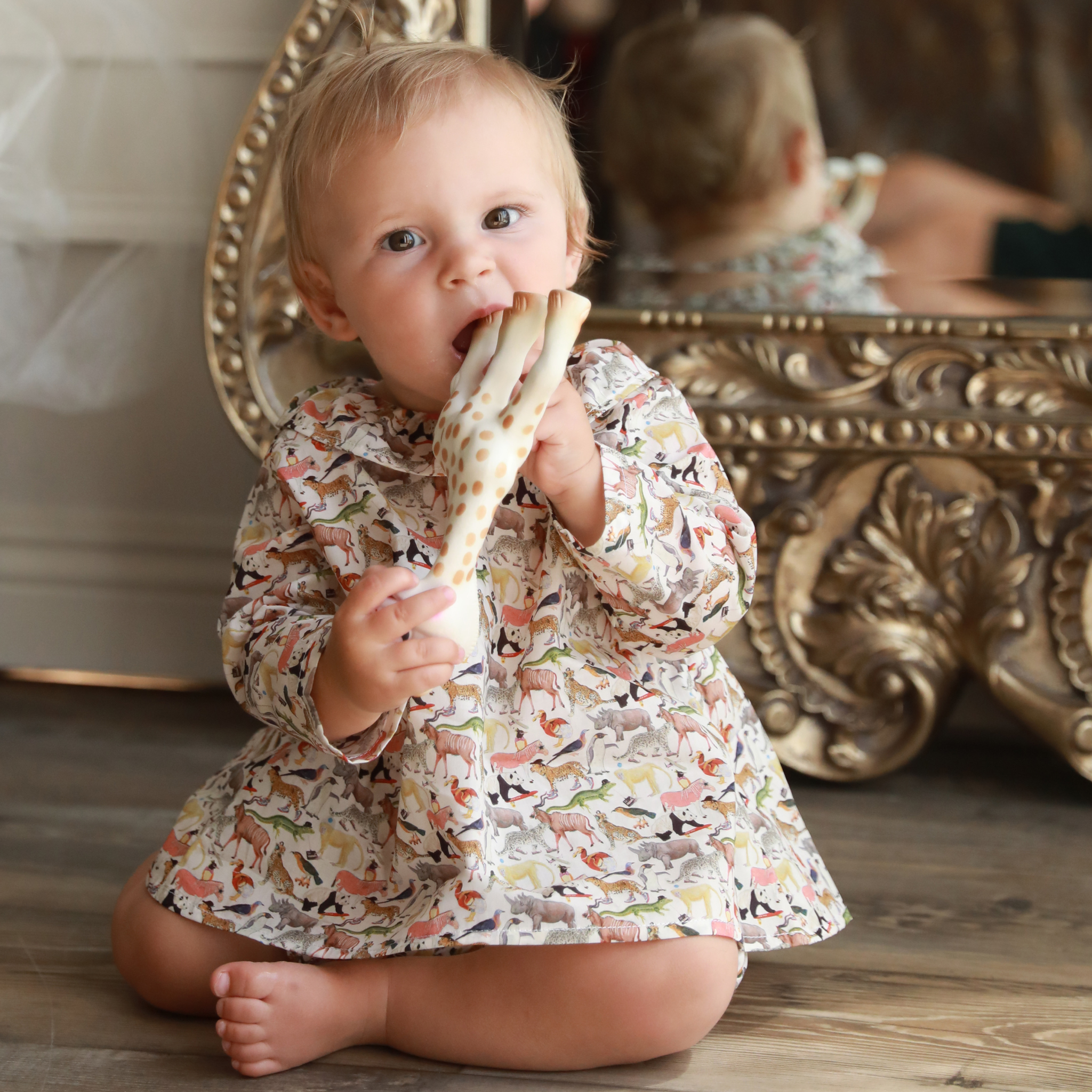 Baby in a patterned blouse sitting on the floor with a decorative mirror in the background.