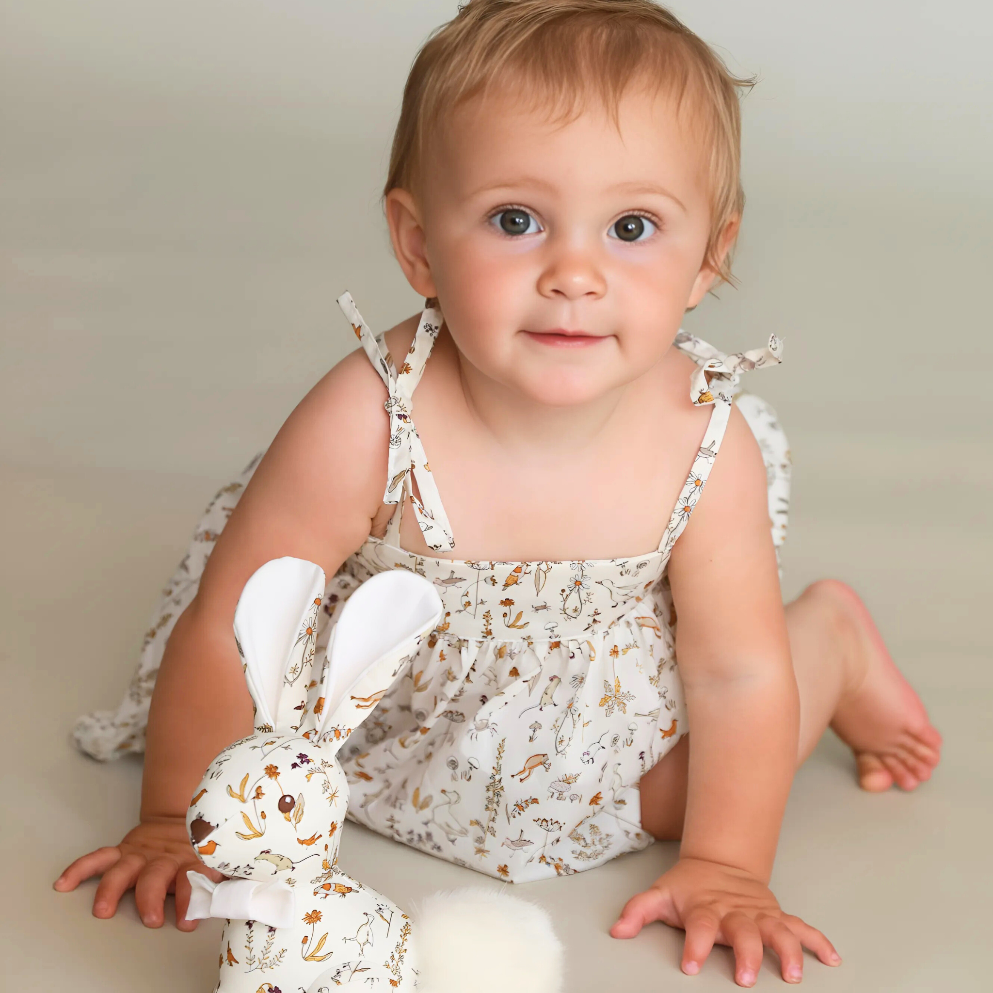 Baby sitting on a beige surface with a floral-patterned outfit and a white toy rabbit.